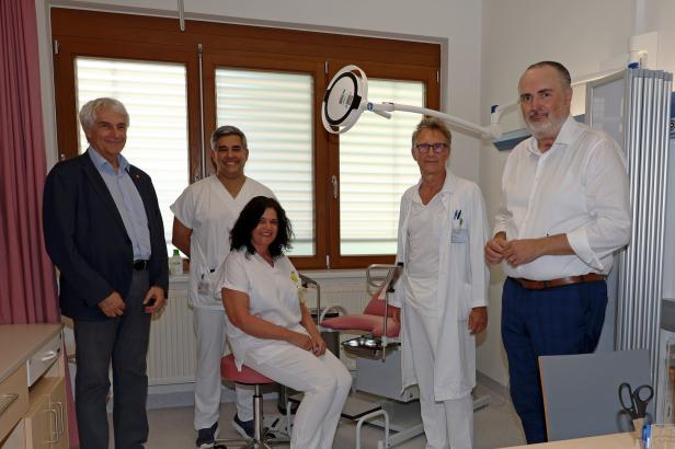 Eine Gruppe von fünf Personen, darunter Ärzte und ein Mann im Anzug, posiert in einem Untersuchungsraum.