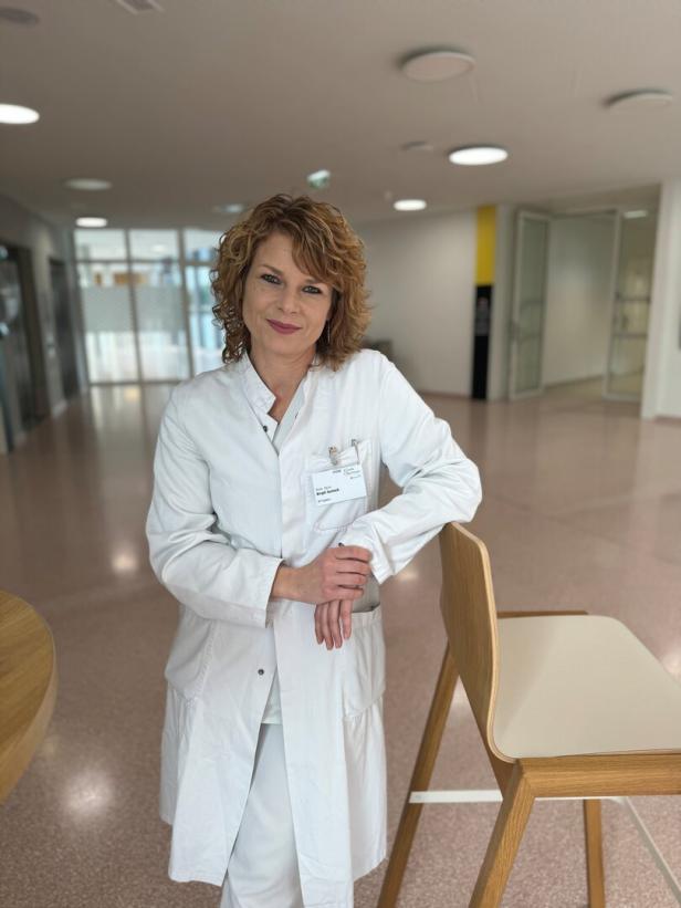 Eine Frau in weißem Kittel steht lächelnd in einem modernen, hellen Flur neben einem Holzstuhl.