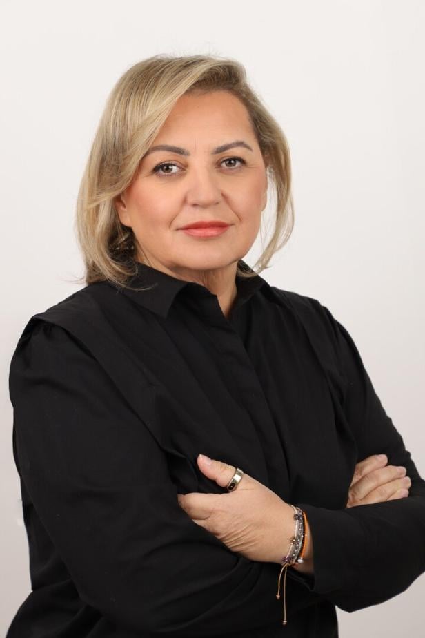 Frau mit blondem Haar und schwarzer Bluse steht mit verschränkten Armen vor hellem Hintergrund.