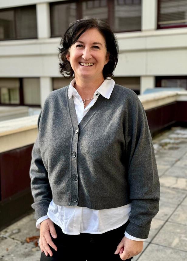 Eine Frau mit dunklen Haaren lächelt und trägt eine graue Strickjacke über einer weißen Bluse im Freien.