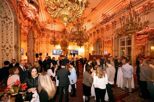 Menschen feiern in einem prunkvollen, festlich beleuchteten Saal mit goldenen Kronleuchtern.