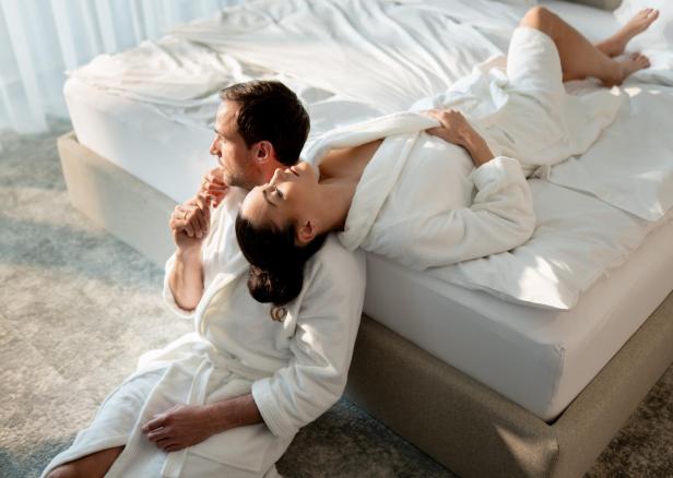 Eine Frau liegt in einem Bademantel auf einem Bett, ein Mann sitzt auf einem Boden, ans Bett gelehnt 