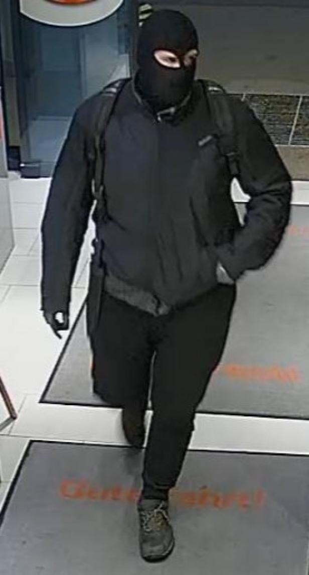 Maskierter Mann in dunkler Kleidung mit Rucksack betritt einen hell erleuchteten Raum.