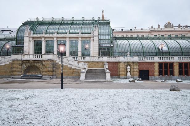 Ein historisches Gewächshaus mit Glasdach und grüner Metallstruktur steht vor leicht verschneitem Rasen.