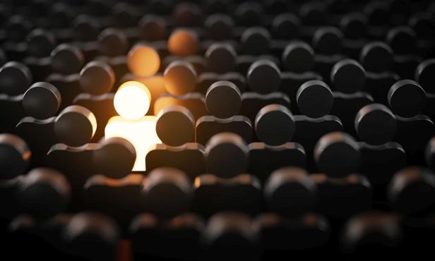 Viele schwarze Figuren stehen dicht beieinander, eine leuchtende Figur hebt sich in der Mitte hervor.