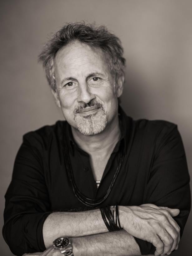 Ein Mann mit grauem Haar und Bart trägt ein dunkles Hemd und mehrere Armbänder, er blickt freundlich in die Kamera.