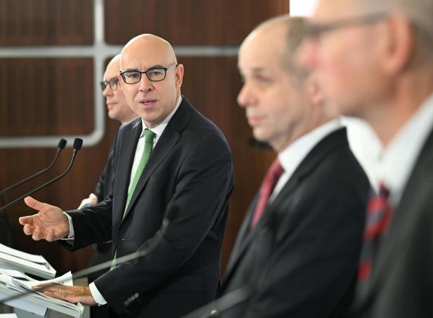 Vier Männer im Anzug stehen nebeneinander an Rednerpulten und sprechen in Mikrofone.