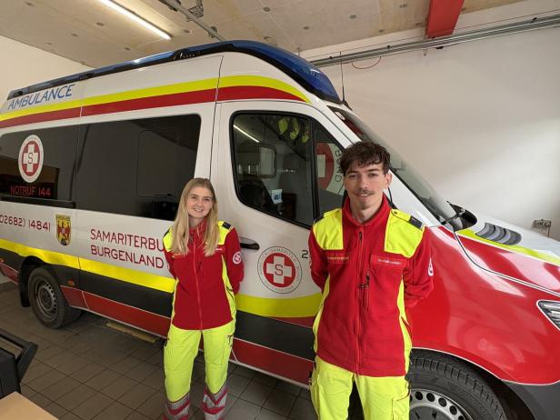 Zwei Rettungskräfte in rot-gelber Uniform stehen vor einem Krankenwagen mit der Aufschrift „Samariterbund Burgenland“.