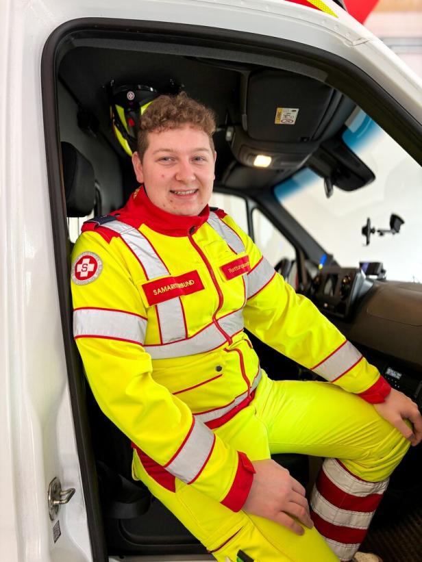 Ein junger Rettungssanitäter in gelber Einsatzkleidung sitzt lächelnd im Fahrerbereich eines Einsatzfahrzeugs.