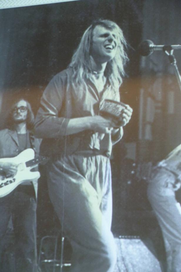 Ein Musiker mit langen Haaren singt und hält ein Instrument, während ein Gitarrist im Hintergrund steht.