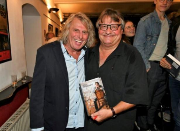 Zwei Männer lächeln, einer hält ein Buch mit einem Porträt auf dem Cover, im Hintergrund weitere Personen.