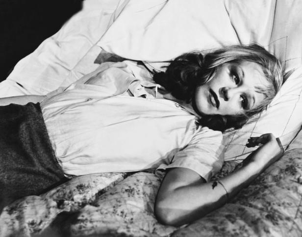 Eine Frau mit welligem Haar liegt nachdenklich auf einem Bett mit gemustertem Bezug und blickt zur Seite.