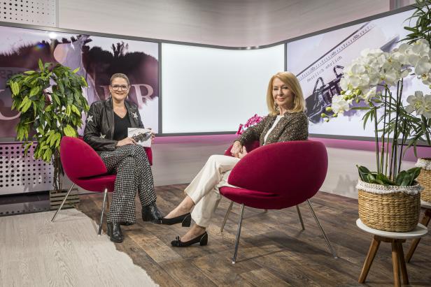 Zwei Frauen sitzen in roten Sesseln in einem modern eingerichteten Studio mit Pflanzen und großen Bildschirmen im Hintergrund.
