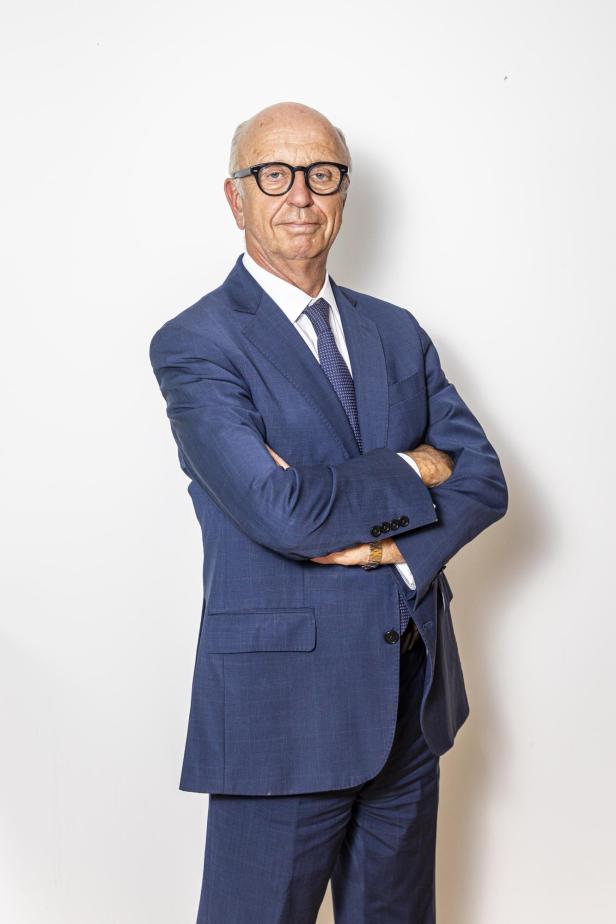 Älterer Mann mit Brille und Glatze trägt blauen Anzug und steht mit verschränkten Armen vor weißem Hintergrund.