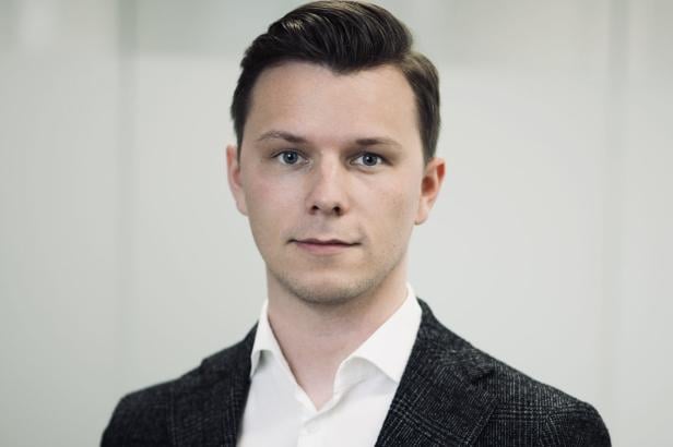 Junger Mann mit kurzen, dunklen Haaren trägt ein weißes Hemd und ein dunkles Sakko, neutraler Hintergrund.