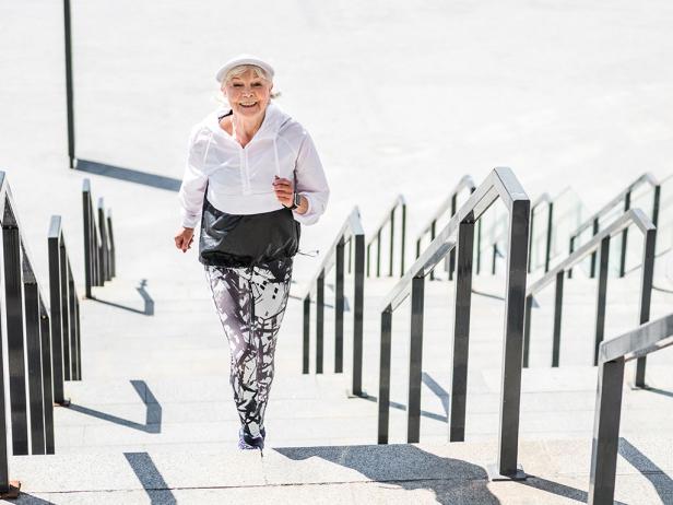 Ältere Frau in Sportkleidung läuft lächelnd eine Treppe im Freien hinauf.