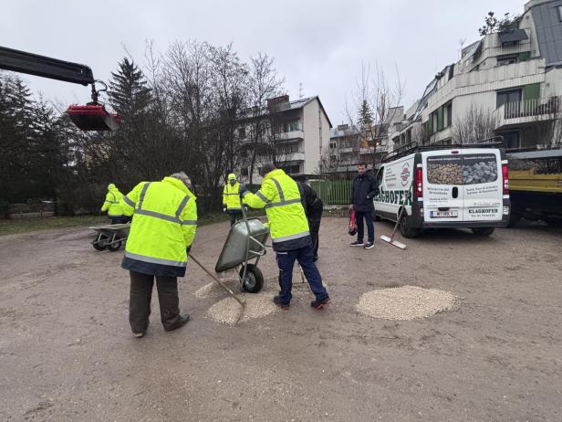 Mehrere Personen füllen Schlaglöcher mit Schotter auf einer Straße vor Wohnhäusern.