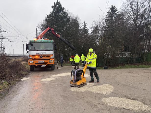 Mehrere Personen füllen Schlaglöcher mit Schotter auf einer Straße vor Wohnhäusern.