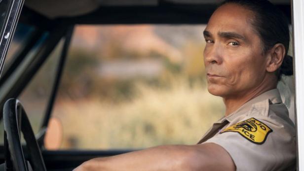 Ein Szene aus "Dark Winds": Ein Mann (Zahn McClarnon) in Uniform mit "Navajo Police"-Abzeichen sitzt im Auto und blickt ernst aus dem Fenster.
