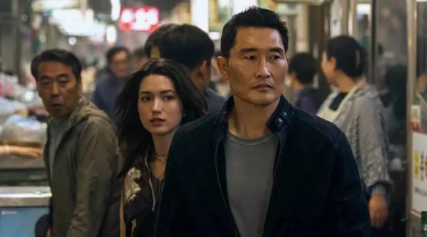 Eine Szene aus "Butterfly": Mehrere Menschen gehen durch einen belebten Marktgang, zwei Personen stehen im Vordergrund und schauen ernst.