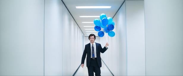 Ein Mann im Anzug geht durch einen weißen Flur und hält einen Strauß blauer Luftballons in der Hand.