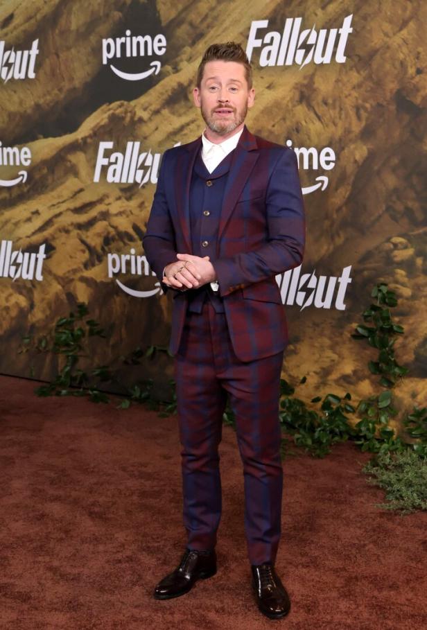 Ein Mann im karierten Anzug steht auf einem braunen Teppich vor einer Wand mit „Fallout“- und „prime“-Logos sowie grünen Pflanzen.