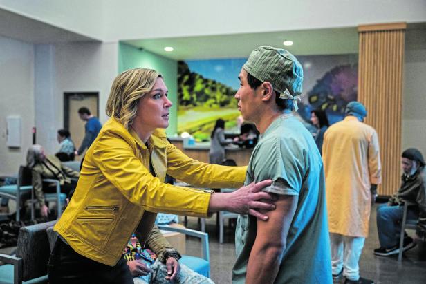 Eine Frau in gelber Jacke hält einen Mann in OP-Kleidung fest und spricht ihn in einem Wartebereich an.