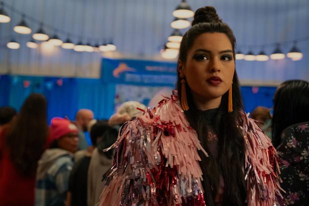 Eine Szene aus "North of North": Eine junge Frau (Anna Lambe) mit langen dunklen Haaren und auffälliger Fransenkleidung steht inmitten einer Menschenmenge in einer Halle.