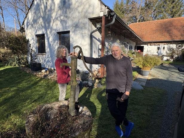 Zwei ältere Personen stehen an einer alten Wasserpumpe im Garten vor einem weißen Haus mit rotem Dach.