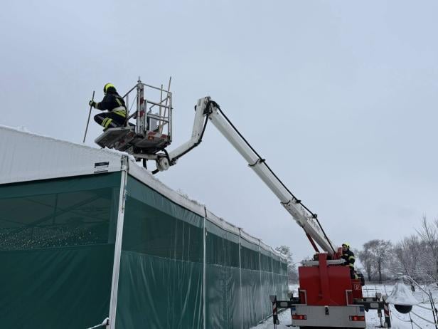 Zwei Feuerwehrleute entfernen mit einer Hubarbeitsbühne Schnee vom Dach eines großen Zeltes.