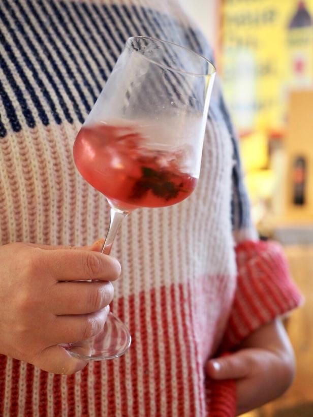 Eine Person hält ein Weinglas mit einem rosafarbenen Getränk und Eiswürfeln vor einem gestreiften Pullover.