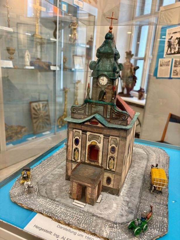 Ein detailreiches Modell einer Kirche mit Uhrturm, umgeben von kleinen Figuren, Kutsche und Fahrzeugen auf gepflasterter Straße.