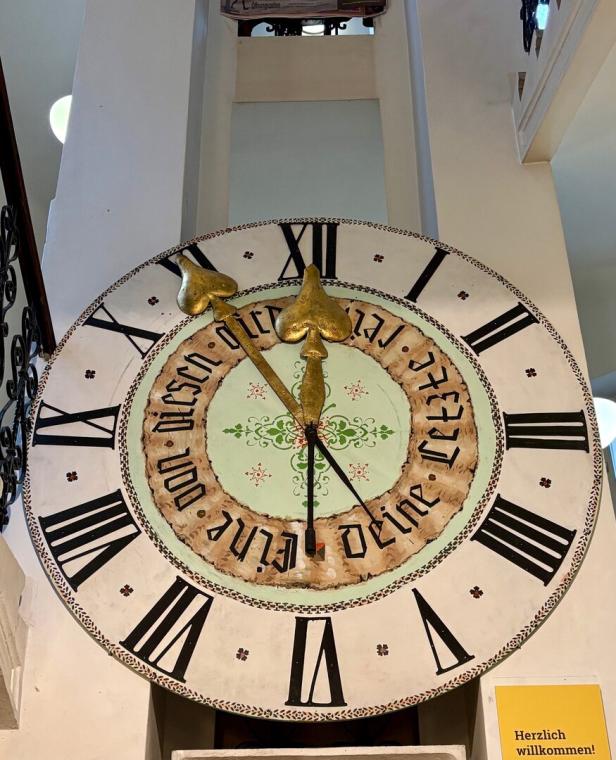 Große Wanduhr mit römischen Ziffern, goldenen Zeigern und einem dekorativen Muster in der Mitte.