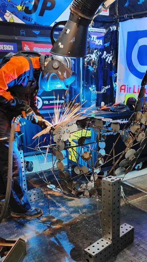 Eine Person mit Schutzkleidung schweißt an einer Metallskulptur, während Funken sprühen.