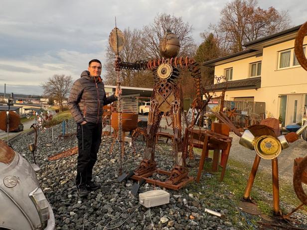Ein Mann steht neben einer großen Skulptur aus Metallteilen im Freien vor einem Gebäude.