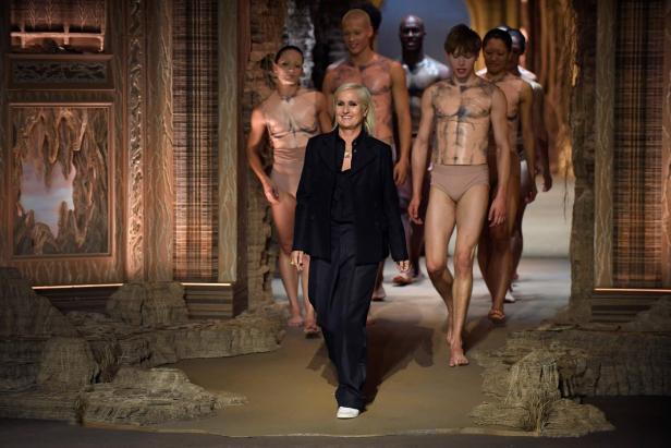 Eine Frau in dunkler Kleidung geht vor mehreren Models in hautfarbenen Outfits auf einem kunstvoll gestalteten Laufsteg.