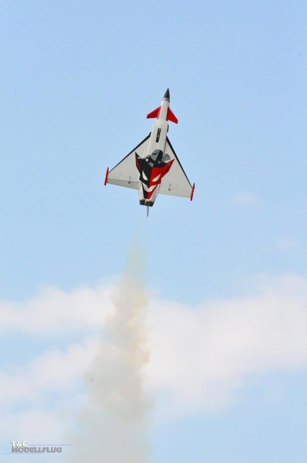 Ein Modellflugzeug mit roter und schwarzer Lackierung steigt senkrecht in den Himmel und hinterlässt eine Rauchspur.