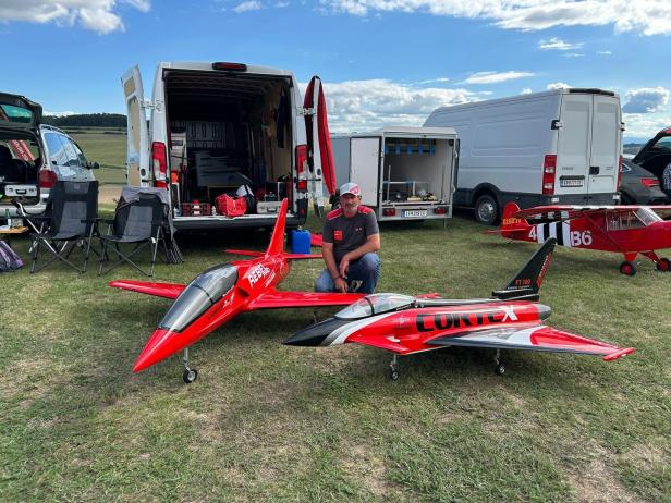 Ein Mann kniet auf einer Wiese neben zwei großen, roten Modellflugzeugen, im Hintergrund stehen geöffnete Transporter.