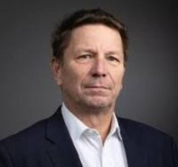 Mann mittleren Alters mit kurzem braunem Haar trägt Anzug und weißes Hemd vor dunklem Hintergrund.