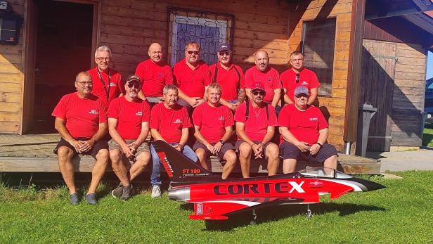 Elf Männer in roten T-Shirts sitzen und stehen vor einer Holzhütte, vor ihnen steht ein großes Modellflugzeug.