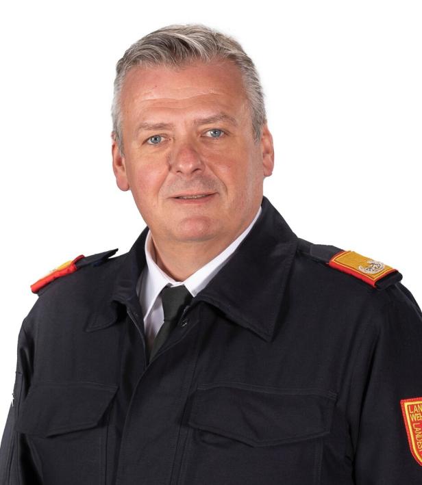 Ein Mann mit grauen Haaren trägt eine dunkelblaue Uniform mit Abzeichen auf den Schultern und am Arm.