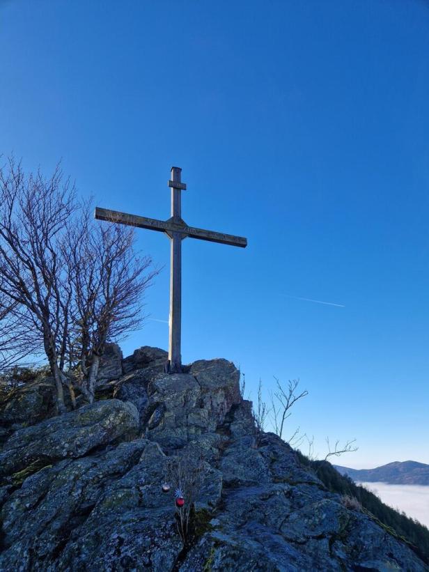 Ein Holzkreuz steht auf einem felsigen Gipfel, daneben kahle Bäume und im Hintergrund blauer Himmel.