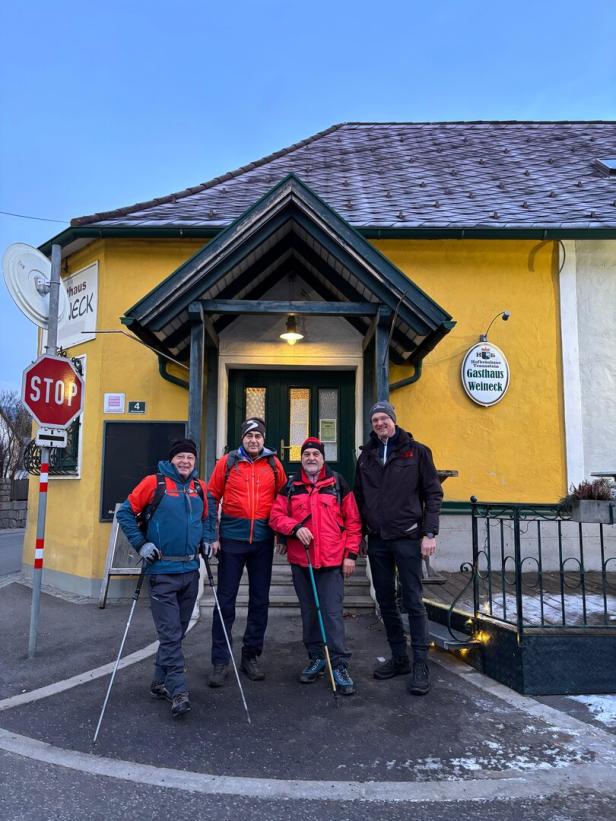 Vier Männer in Outdoor-Kleidung mit Wanderstöcken stehen vor einem gelben Gebäude mit der Aufschrift „Gasthaus Weineck“.