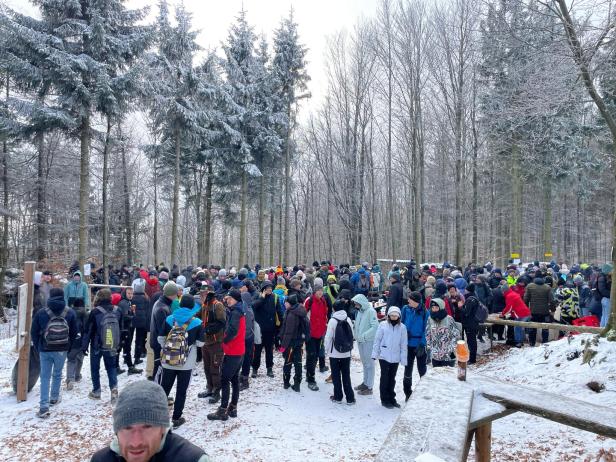 Viele Menschen mit Winterkleidung versammeln sich auf einem verschneiten Waldweg zwischen schneebedeckten Bäumen.