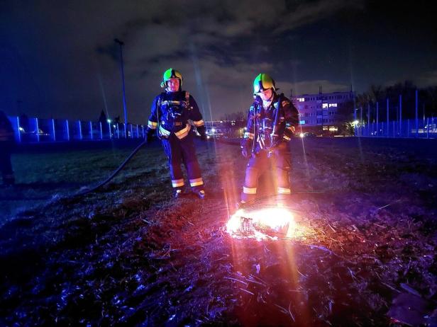 Zwei Feuerwehrleute in Schutzkleidung stehen nachts auf einer Wiese und löschen ein kleines Feuer am Boden.