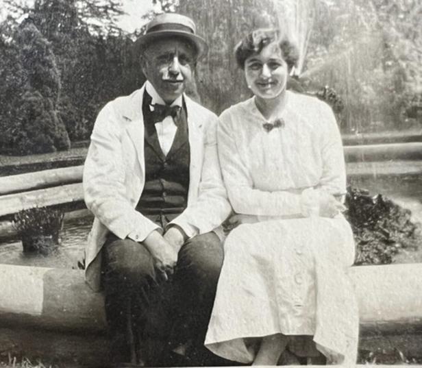 Ein Mann mit Hut und eine Frau sitzen vor einem Brunnen im Freien.