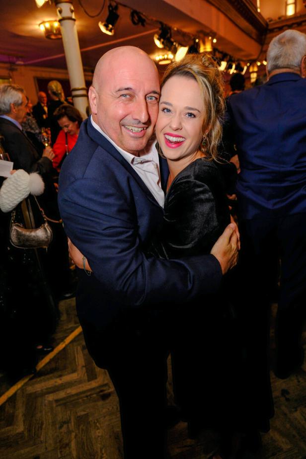 Ein Mann und eine Frau umarmen sich lachend auf einer festlichen Veranstaltung mit vielen Menschen im Hintergrund.