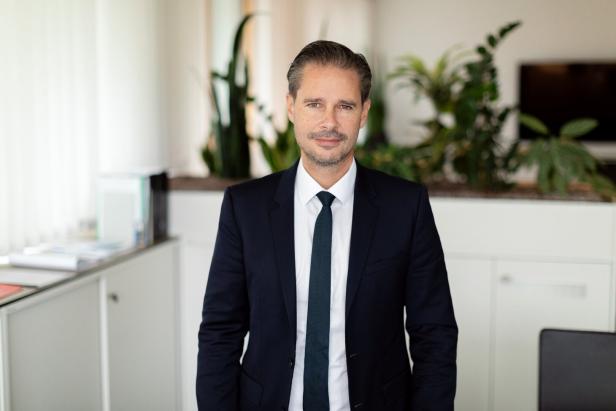 Ein Mann im Anzug steht in einem modernen Büro mit Pflanzen und weißen Möbeln im Hintergrund.