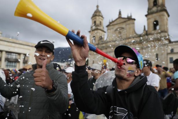 Zwei Männer feiern mit einer bunten Tröte und Daumen hoch vor einer historischen Kirche, umgeben von Konfetti und Menschenmenge.