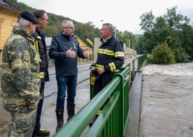 Vier Männer in Uniformen stehen auf einer Brücke und besprechen die Hochwassersituation am reißenden Fluss.